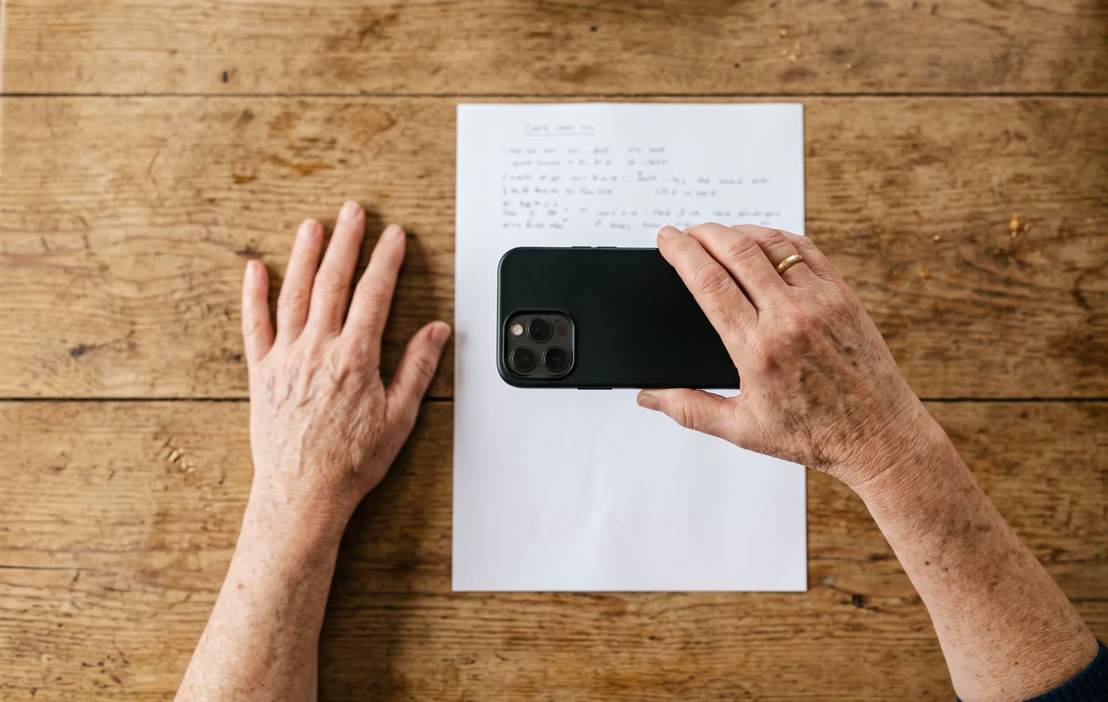 Draufsicht auf einen Holztisch: Zwei Hände einer älteren Person, eine hält ein Smartphone über ein Blatt Papier, um es zu fotografieren